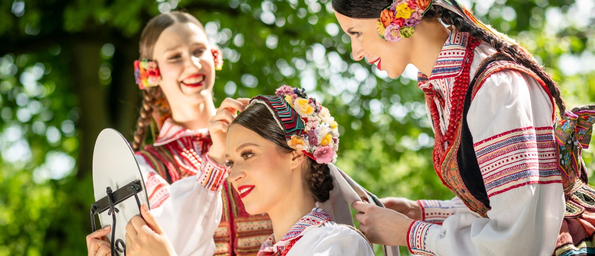 Dwie artystki Zespołu Mazowsze w kostiumach krzczonowskich stroją głowę artystki siedzącej przed lusterkiem.