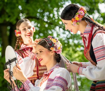 Dwie artystki Zespołu Mazowsze w kostiumach krzczonowskich stroją głowę artystki siedzącej przed lusterkiem.