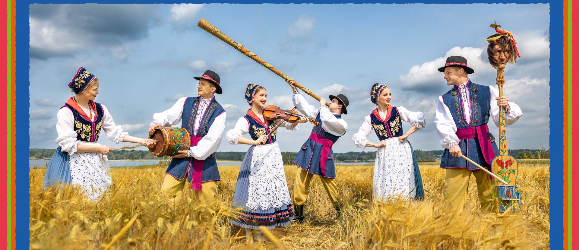 Artyści Zespołu Mazowsze z instrumentami kaszubskimi i ubrani w kostiumy kaszubskie pozują w polu ze zbożem przy jeziorze..