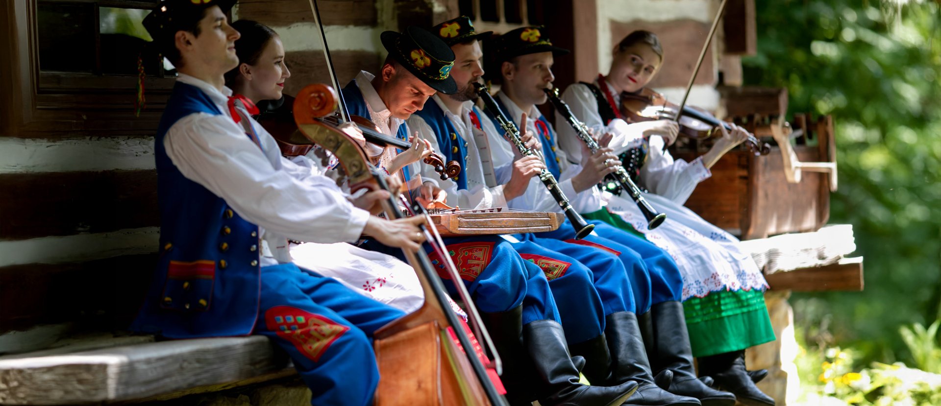 Członkowie Kapeli Trampolka, z instrumentami w rękach, siedzą na ławce przy chacie