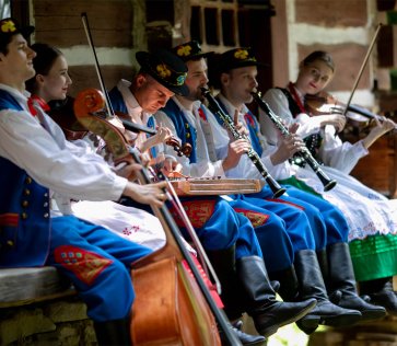 Członkowie Kapeli Trampolka, z instrumentami w rękach, siedzą na ławce przy chacie