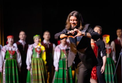Tomasz Szczepanik podczas koncertu na scenie, w tle widoczny chór "Mazowsza" w kostiumach kurpiowskich