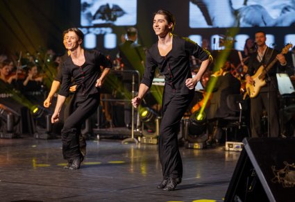 Tancerze ubrani na czarno na scenie podczas układu, w tle widoczna orkiestra