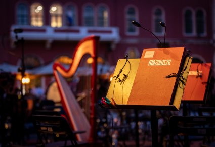 Widać pulpity orkiestrowe ustawione na scenie podczas koncertu na świeżym powietrzu. Na pierwszym planie znajduje się brązowa teczka z napisem „Mazowsze”, W tle widać instrumenty, w tym harfę, oraz zarys budynku z arkadowymi oknami