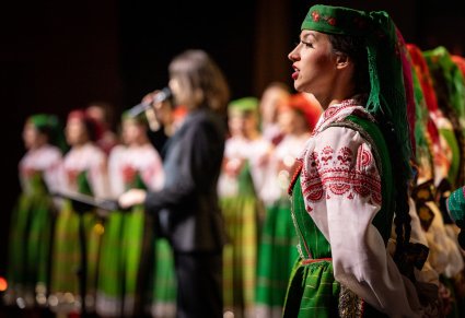 Zbliżenie na chórzystkę w kostiumie kurpiowskim, w tle reszta chóru oraz wokalista zespołu Pectus