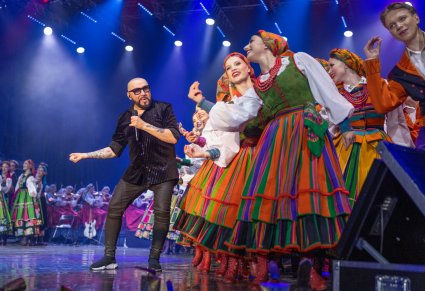Na pierwszym planie na scenie Solista zespołu Kombii wraz z Artystami w kostiumach łowickich w trakcie wspólnego występu na scenie, w tle p część Artystów Chóru i Orkiestra w kostiumach wilanowskich