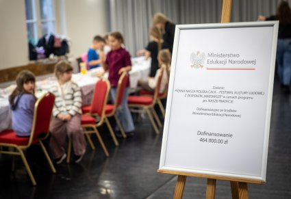 Zbliżenie na tablicę informacyjną o dofinansowaniu, w tle widoczne dzieci podczas warsztatów