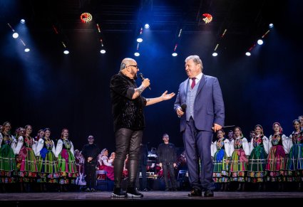 Na środku sceny Dyrektor zespołu "Mazowsze" wraz z Solistą zespołu Kombii. Za nimi Artyści Chóru w kostiumach łowickich wraz z Orkiestrą w kostiumach wilanowskich