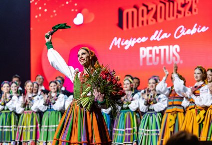 Tancerka w kostiumie sannickim z kwiatami na scenie, w tle zespół Mazowsze