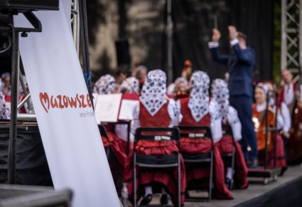 Na pierwszym planie rollup z logo Mazowsze Serce Polski w tle Dyrygent wraz z Orkiestrą w kostiumach wilanowskich