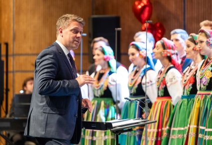 Zbliżenie na Dyrygenta w ciemnym garniturze, zwróconego w stronę Artystów Chóru w kostiumach łowickich