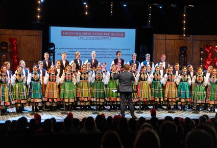 Na scenie Artyści Chóru w kostiumach łowickich podczas śpiewu, przed nimi, zwrócony w ich stronę Dyrygent w ciemnym garniturze. Widoczna część Publiczności