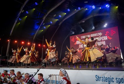 Na scenie Artyści w kontuszach wykonujący choreografię taneczną, niżej pod sceną część orkiestry