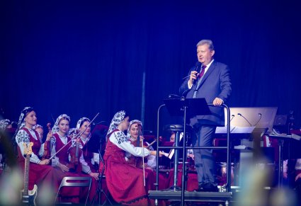 Dyrygent- Dyrektor zespołu "Mazowsze" przemawiający do Publiczności, obok niego, siedząca Orkiestra w kostiumach wilanowskich
