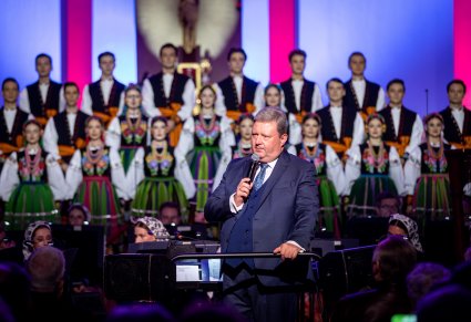 Dyrektor przemawiający przed koncertem, chór i orkiestra widoczne w tle