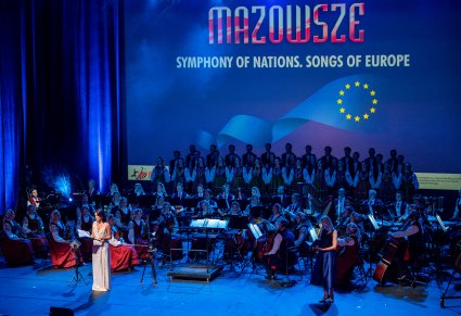 Widok na scenę z Artystami i konferansjerem. Tłem sceny jest ekran na którym widoczny jest napis MAZOWSZE SYMPHONY OF NATIONS, SONGS OF EUROPE
