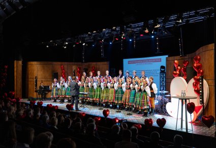 Na scenie Artyści Chóru w kostiumach łowickich w trakcie śpiewu, przed nimi Dyrygent zwrócony w ich stronę, a z boku sceny Akompaniator grający na instrumencie. Widoczna część Publiczności