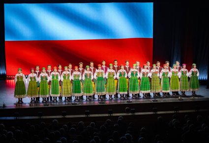 Artyści chóru w kostiumach kurpiowskich podczas śpiewu w tle flaga Polski