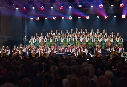 Na oświetlonej scenie Artyści Chóru w kostiumach łowickich, poniżej Artyści Orkiestry w kostiumach wilanowskich wraz z Dyrygentem