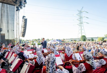 Widok na Artystów Orkiestry w kostiumach wilanowskich przed nimi Dyrygent w arniturze trzymający w ręku batutę, za nimi widoczna część Publiczności koncertowej