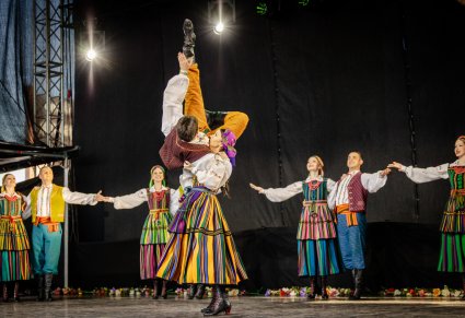 Na środku sceny para Artystów Baletu w kostiumach opoczyńskich wykonująca partnerowanie przy choreografii tanecznej, za nimi reszta część Artystów stojąca w parach