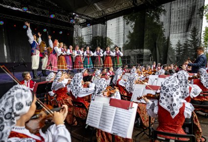 Na pierwszym planie siedzące tyłem skrzypaczki i dalsza część orkiestry z widocznym dyrygentem. Na scenie Artyści w kostiumach limanowskich