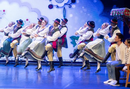 Grupa tancerzy w kostiumach kaszubskich wchodząca na scenę, obok aktorzy siedzący na ławce obserwujący taniec