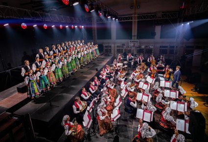 Boczny widok na scenę na której znajdują się Artyści Chóru w kostiumach łowickich, Artyści Orkiestry w kostiumach wilanowskich oraz Dyrygent trzymający w ręku batutę