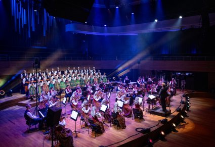 Widok na scenę na ktorej znajduje się Dyrektor zespołu "Mazowsze", dyrygujący koncert, zwrócony w stronę Orkiestry w kostiumach wilanowskich i Artystów Chóru w kostiumach łowickich