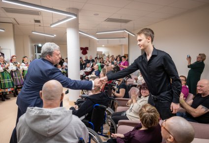Dyrektor zespołu "Mazowsze" podający dłoń Pacjentowi Kliniki "Budzik" dla Dorosłych, w tle reszta część Pacjentów Kliniki i część Artystów Chóru w kostiumach łowickich