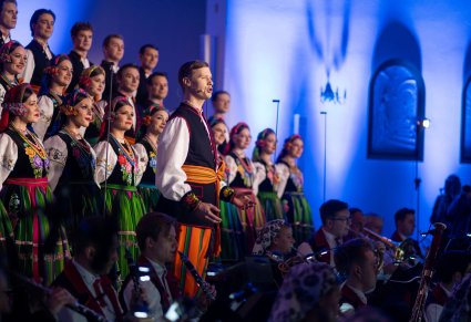 Na pierwszym planie śpiewający solista w kostiumie łowickim, w tle część Artystów Chóru, niżej część Orkiestry w kostiumach wilanowskich
