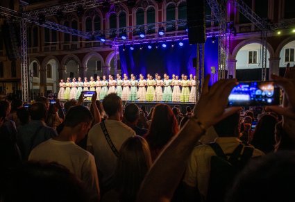 Artyści na scenie w stylowych kostiumach XIX w., widoczna publiczność, która robi zdjęcia telefonami komórkowymi