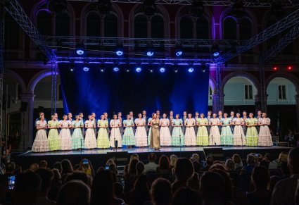 Artyści na scenie w stylowych kostiumach XIX w. a przed nimi Solistka w długiej, złotej sukience. Widoczna część publiczności