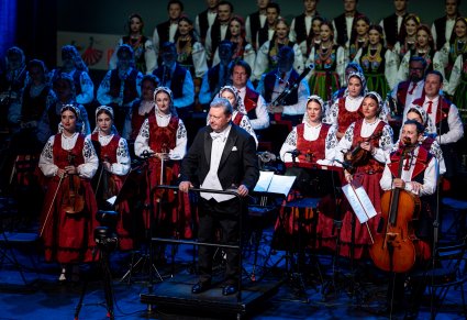 Na pierwszym planie Dyrektor Jacek Boniecki jako Dyrygent za nim orkiestra a na trzecim planie chór w rzędach w kostiumach łowickich