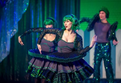 Zbliżenie na dwie Artystki odgrywające role postaci kół w zielonych kostiumach wykonujące choreografię taneczną, za nimi jeden Artysta również w zielonym kostiumie