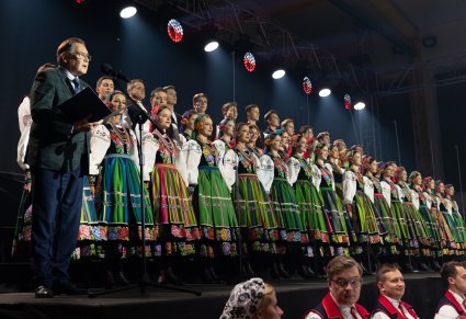 Na pierwszym planie były Kierownik Chóru przemawiający do mikrofonie, w tym samym rzędzie Artyści Chóru w kostiumach łowickich