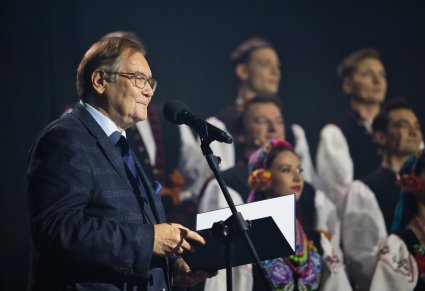 Zbliżenie na prowadzącego koncert, trzymającego w ręku granatową teczkę, w tle widoczna część Artystów Chóru w kostiumach łowickich