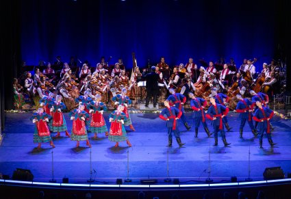 Na scenie grupa Artystów w kostiumach kujawskich w trakcie wykonywania choreografii tanecznej, w tle widoczna Orkiestra w kostiumach wilanowskich