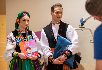 Para Artystów w kostiumach łowickich trzymająca w rękach prezenty dla Dzieci z Kliniki "Budzik". Książeczkę "Wielkie marzenie małego Tadzia" i poduszkę na której widoczna jest wyhaftowana choinka