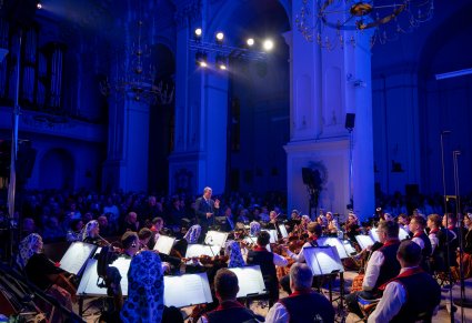 Na pierwszym planie Dyrektor zespołu "Mazowsze" dyrygujący koncert, za nim publiczność, przed nim Orkiestra w kostiumach wilanowskich