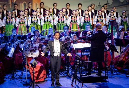 Na pierwszym planie solista ubrany w smoking za nim Dyrektor Jacek Boniecki Dyrygujący koncert, następnie Orkiestra i chór ustawiony w rzędy w kostiumach łowickich