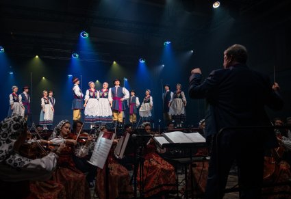 Na pierwszym planie Dyrygent zwrócony w stronę Orkiestry i Artystów, na scenie Artyści w kostiumach kaszubskich