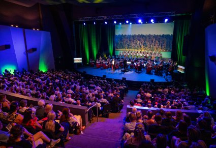 Widok na scenę z widoczną publicznością. Na scenie widoczny solista w smokingu, Dyrektor Jacek Boniecki dyrygujący koncert, Orkiestra i chór ustawiony w rzędy w kostiumach łowickich