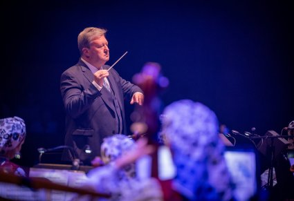 Zbliżenie na Dyrektora zespołu "Mazowsze" , który trzyma w ręku batutę i jest wrócony w stronę Orkiestry i Chóru