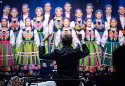 Zbliżenie na dyrygenta podczas koncertu, w tle widoczny chór w kostiumach łowickich