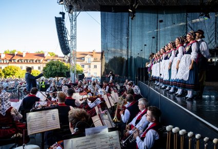 Na pierwszym planie orkiestra a z boku na scenie Artyści stojący w rzędzie w kostiumach kaszubskich