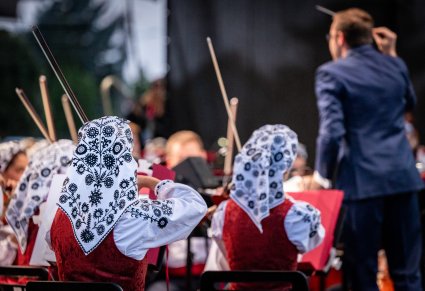 Widok z tyłu orkiestry, zbliżenie na chusty z kostiumu wianowskiego skrzypaczek, w tle dyrygent
