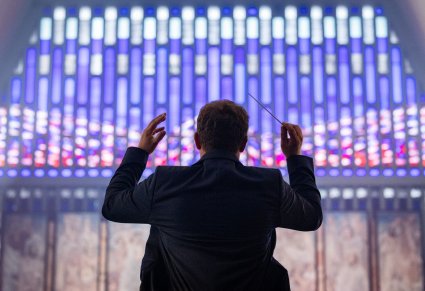 Sylwetka Dyrygenta, trzymająca w górze batutę, zwrócona w stronę Ołtarza