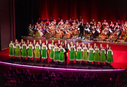 Widok na całą scenę. Na scenie na pierwszym planie dwie solistki za nimi Artyści Chóru w kostiumach kurpiowskich, na drugim planie Orkiestra w kostiumach wilanowskich