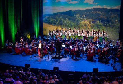 Widok na scenę z widoczną publicznością. Na scenie Solista w smokingu, Dyrektor Jacek Boniecki dyrygujący koncert, Orkiestra a za nią chór ustawiony w rzędy w kostiumach łowickich